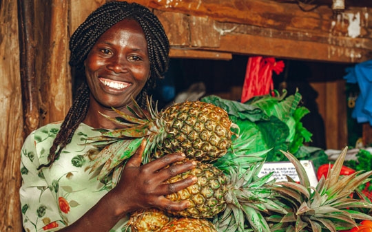 Woman holding pineapples standing next to a fruits and vegetables kiosk