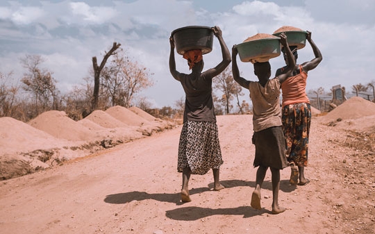 A boy and two women walking holding a bowl on top of their head 